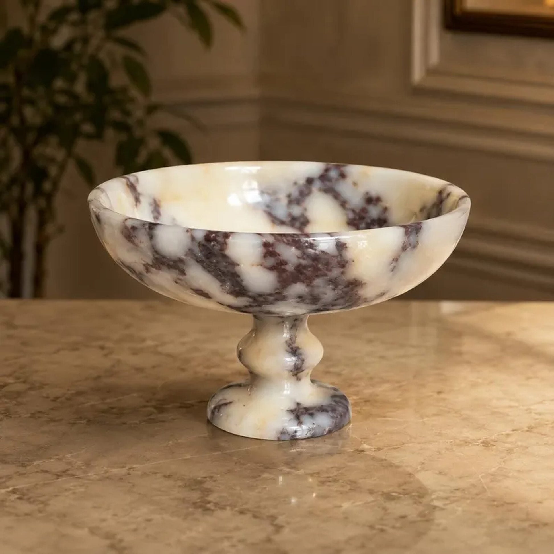 A marble pedestal bowl with a cream and white base and dark brown veining, set on a polished marble countertop in an indoor setting.