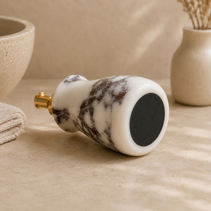 A marble massage roller with a white and dark brown marbled pattern, featuring a gold metal button on one end and a black textured surface on the other, resting on a beige stone surface.