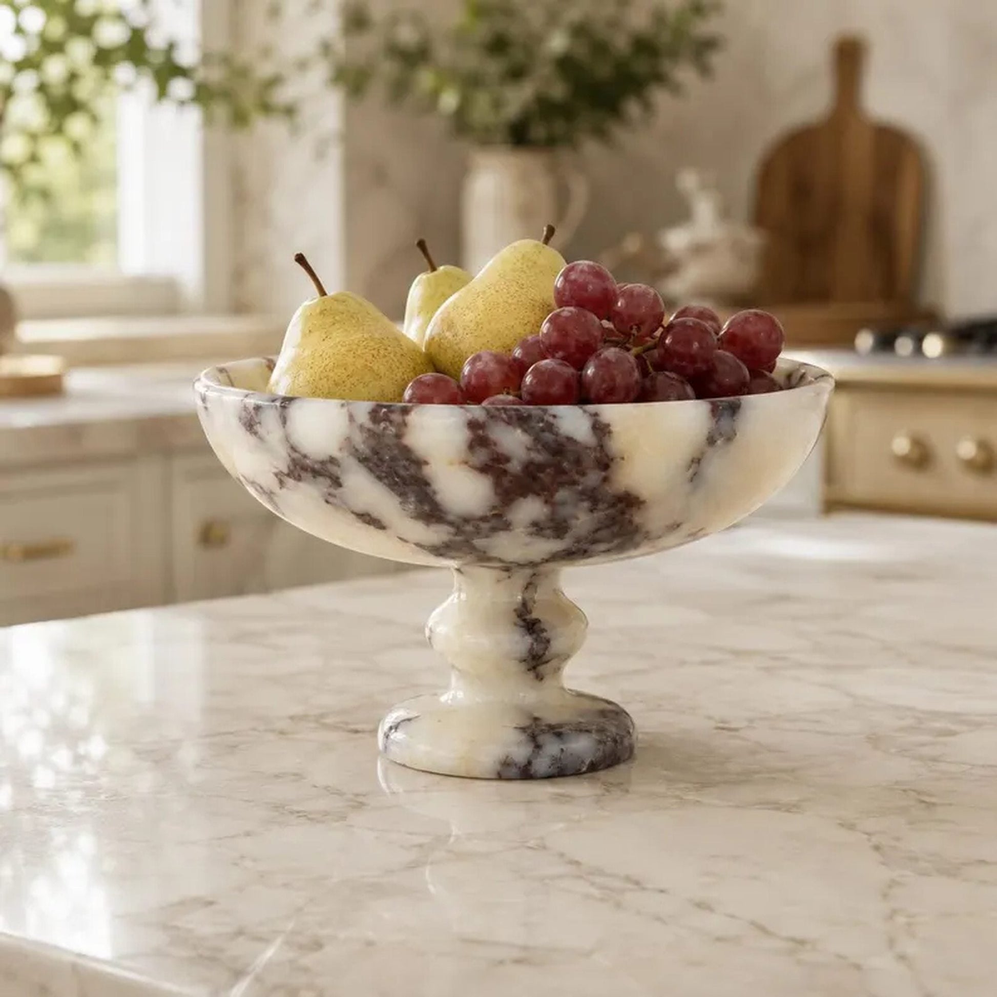 A clear glass pedestal bowl filled with mixed fruit, set on a white pedestal in a bright, modern kitchen interior.