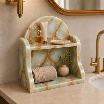 A marble bathroom organizer with a two-tier shelf, styled on a beige marble countertop with a gold faucet, a rolled towel, a small plant, and toiletries.
