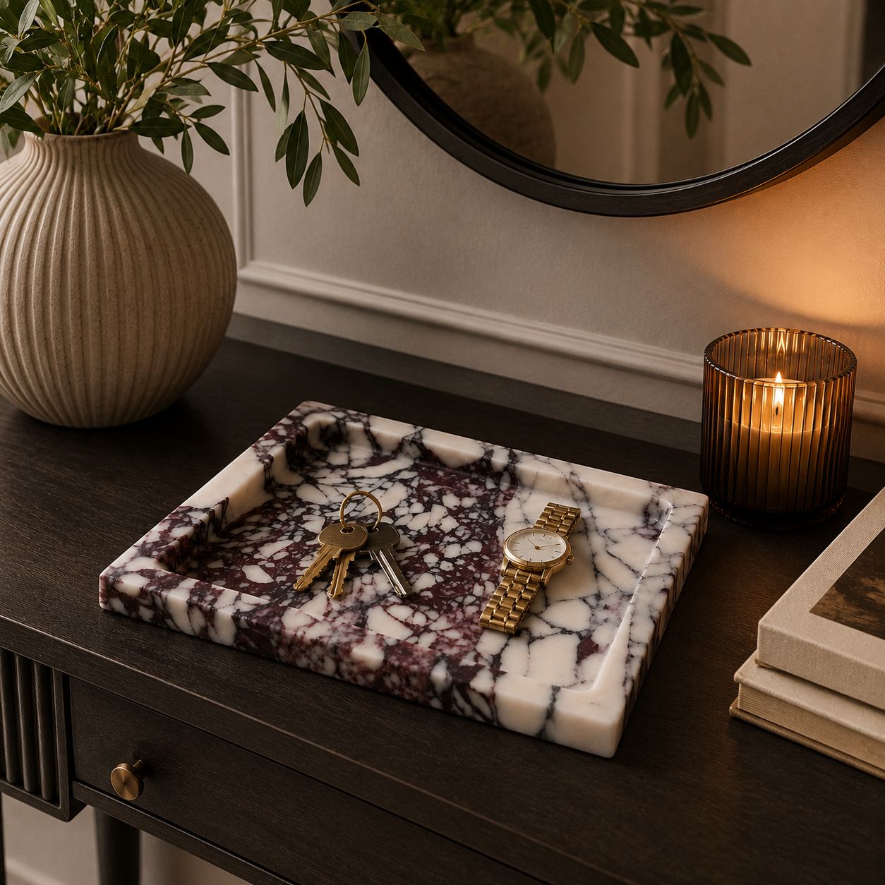 A rectangular marble key tray with a white and dark purple-brown veined pattern sits on a dark wooden console table, styled with keys, a gold watch, a lit candle, a vase with greenery, and a round mirror in the background.