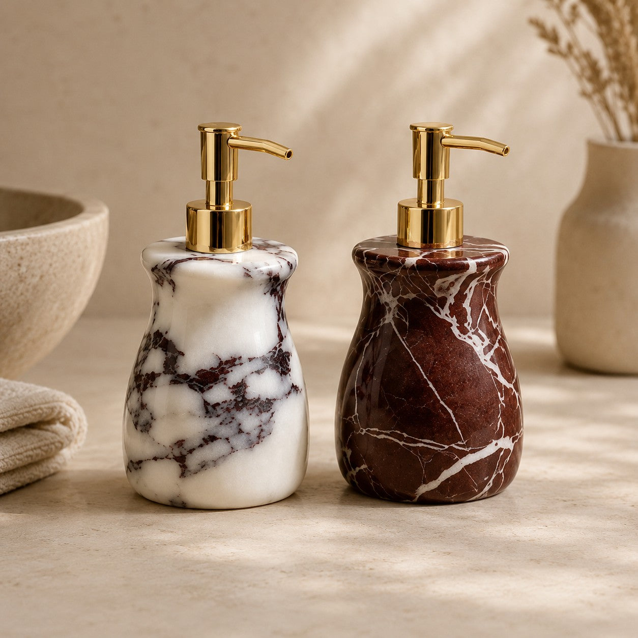Two marble soap or lotion dispensers with gold pump heads, one in white marble with dark veining and one in dark red/burgundy marble with white veining, set on a beige surface with neutral decor in the background.