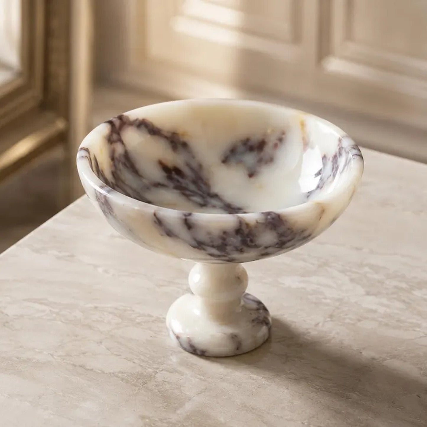 A small marble pedestal bowl with a cream and white base and dark brown veining, set on a light marble tabletop in an elegant interior setting.