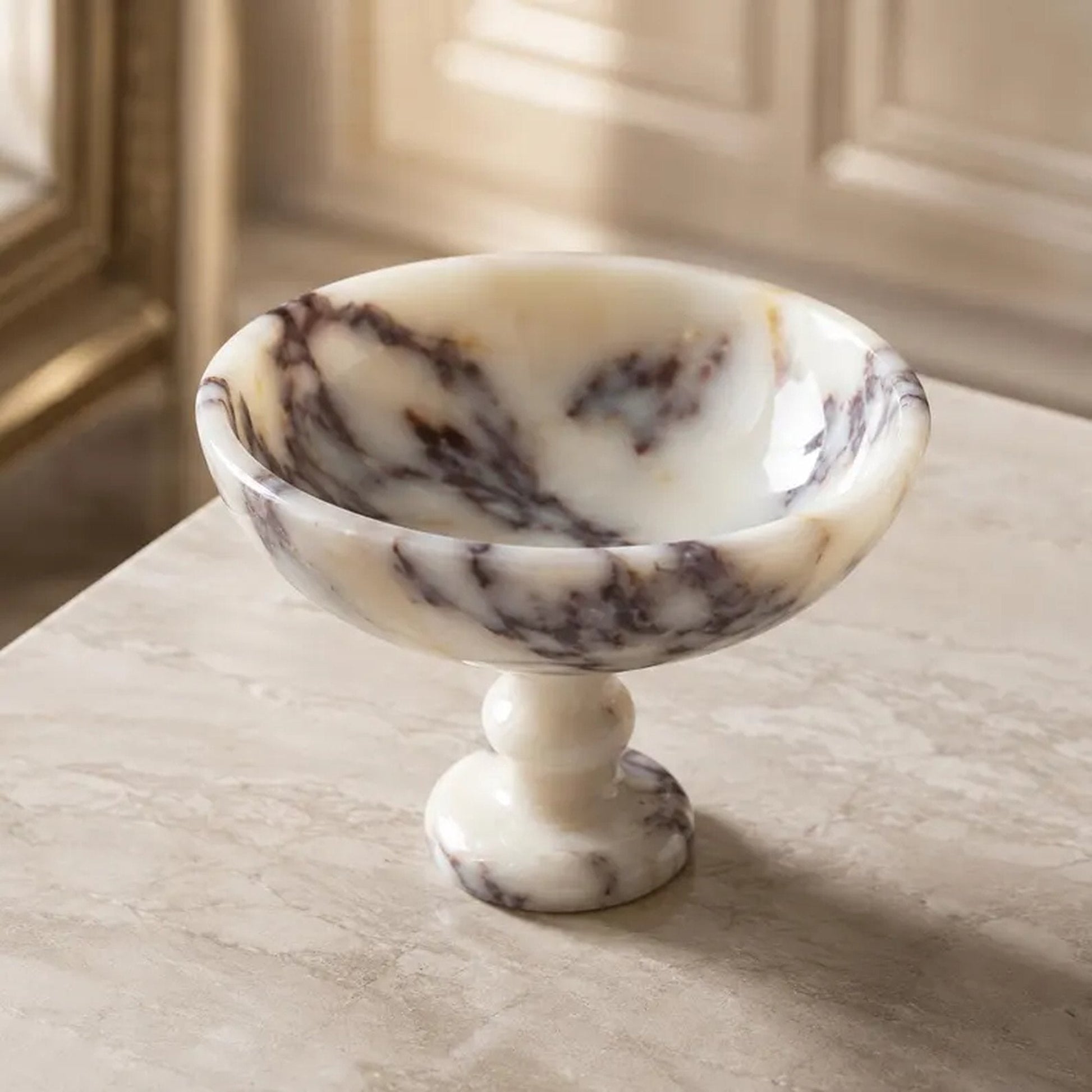 A small marble pedestal bowl with a cream and white base and dark brown veining, set on a light marble tabletop in an elegant interior setting.