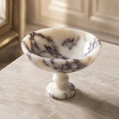 A small marble pedestal bowl with a cream and white base and dark brown veining, set on a light marble tabletop in an elegant interior setting.