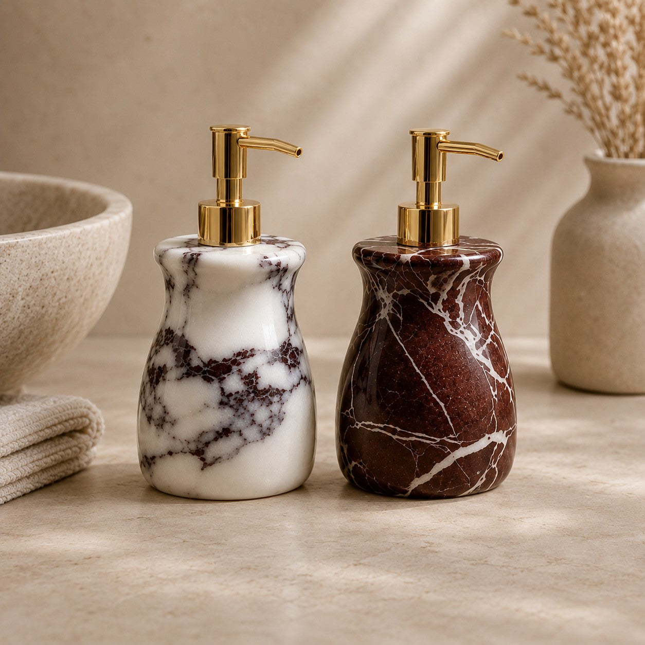 Two marble soap dispensers with gold pumps, one white with dark gray veining and one dark brown with white veining, displayed on a beige stone surface with neutral decorative props.