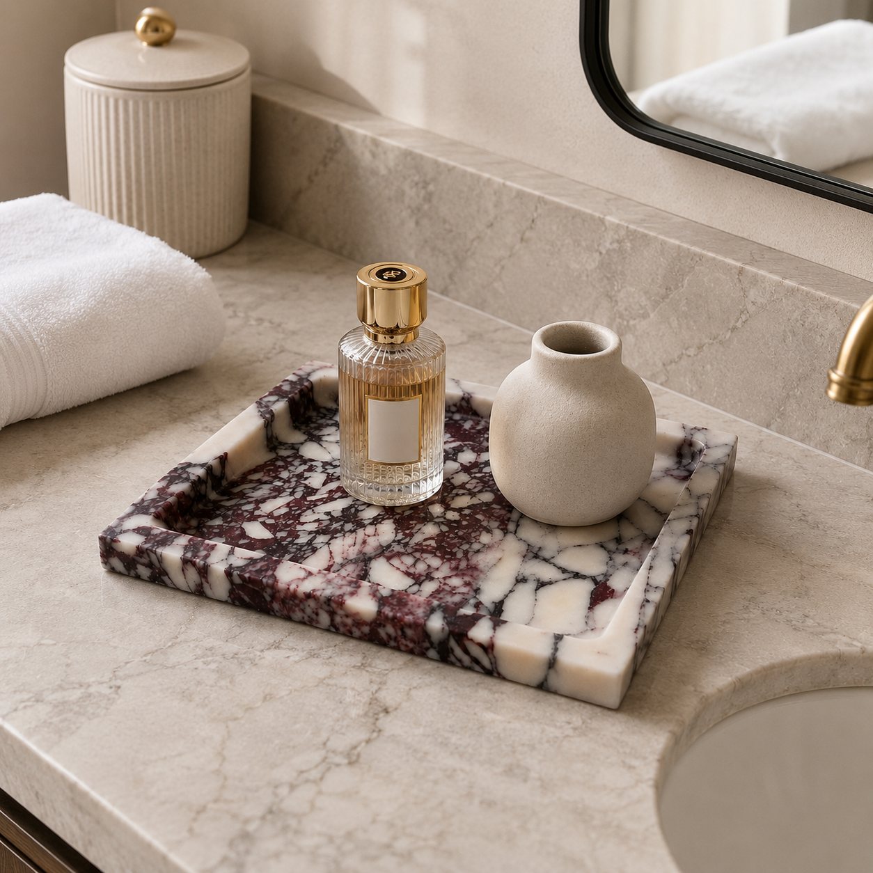 A rectangular marble bathroom tray with a dark veined pattern sits on a marble countertop beside a sink, holding a perfume bottle and a small vase. A folded white towel and a ribbed cream canister are visible in the background.