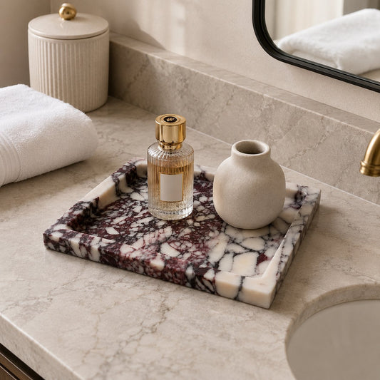 A rectangular marble bathroom tray with a dark veined pattern sits on a marble countertop beside a sink, holding a perfume bottle and a small vase. A folded white towel and a ribbed cream canister are visible in the background.