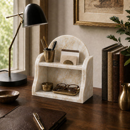 A cream-colored marble desk organizer with two shelves, styled on a dark wooden desk next to a black desk lamp, books, and a plant.