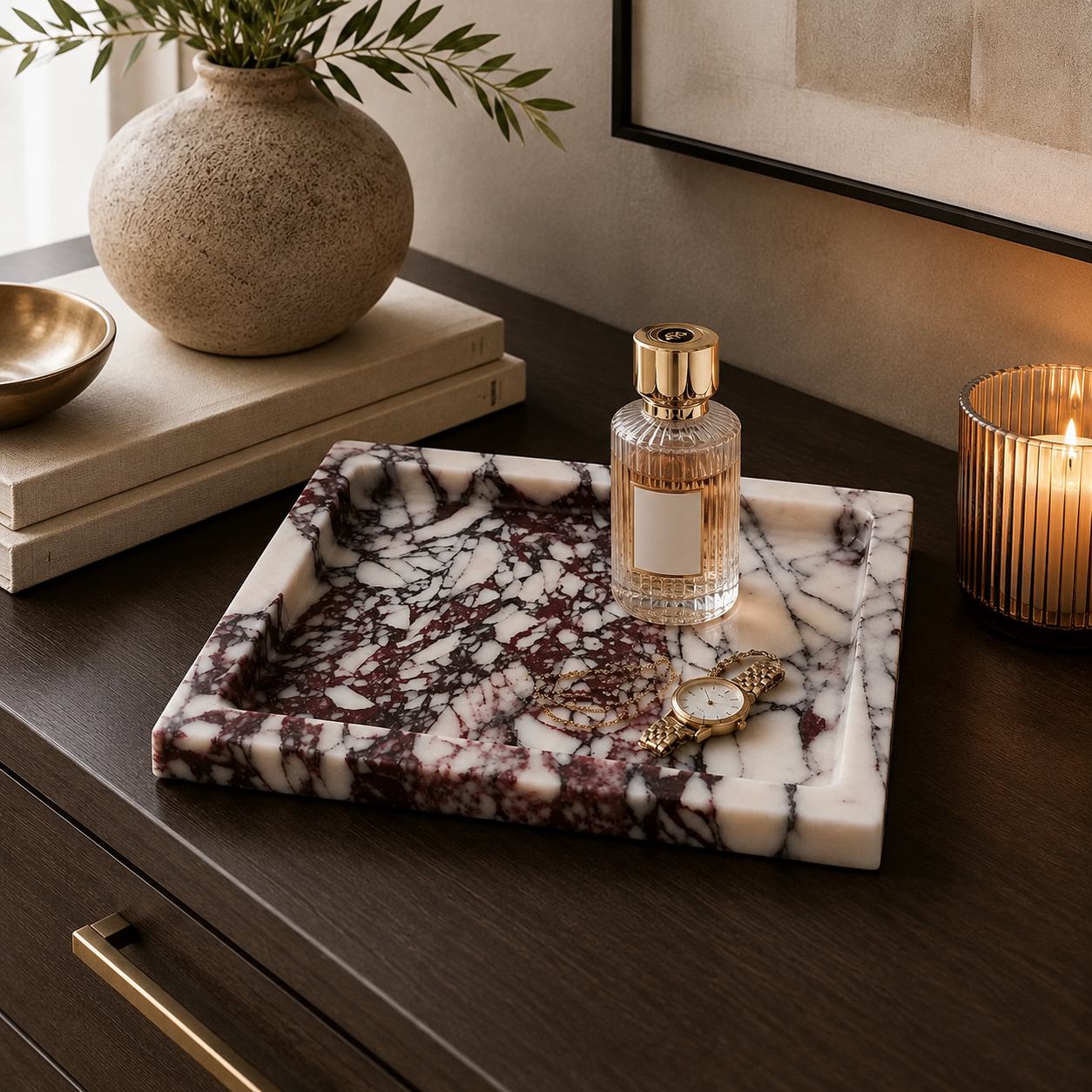 A rectangular marble tray with a white base and dark burgundy veining sits on a dark wood table, styled with a perfume bottle, watch, and gold accessories. In the background, a ceramic vase with greenery and a lit candle are visible.