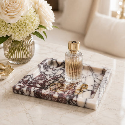 A rectangular marble tray with a white, gray, and burgundy veined pattern sits on a light marble table. A ribbed clear glass perfume bottle with a gold cap rests on the tray, alongside a few gold rings.
