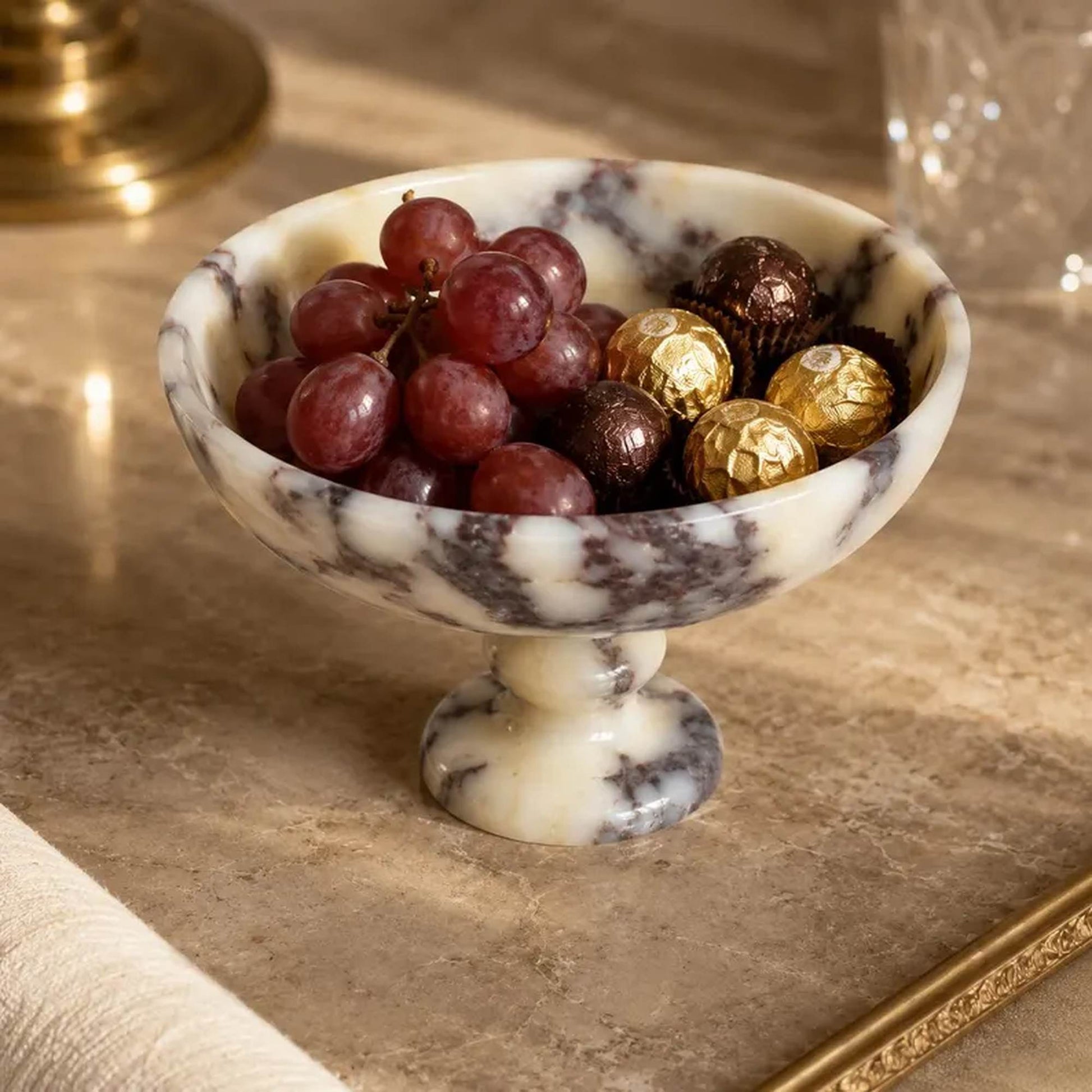 A marble pedestal bowl filled with red grapes and wrapped chocolates, set on a beige stone surface with a blurred warm-toned background.