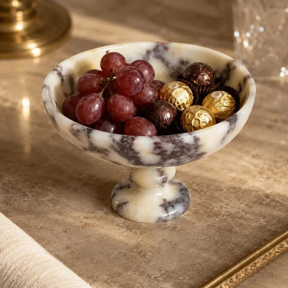 A marble pedestal bowl filled with red grapes and wrapped chocolates, set on a beige stone surface with a blurred warm-toned background.
