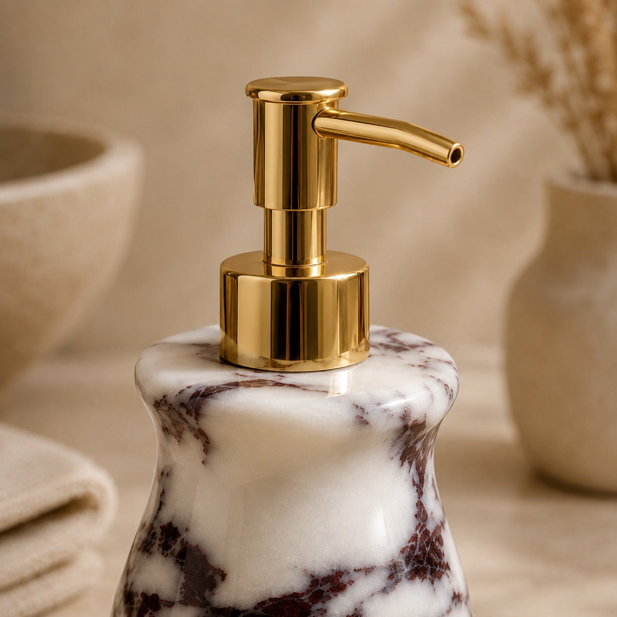 A close-up of a bathroom or vanity pump bottle with a polished gold-tone pump and a white marble base featuring dark brown veining, set against a warm beige background with softly blurred decorative props.