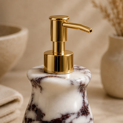 A close-up of a bathroom or vanity pump bottle with a polished gold-tone pump and a white marble base featuring dark brown veining, set against a warm beige background with softly blurred decorative props.