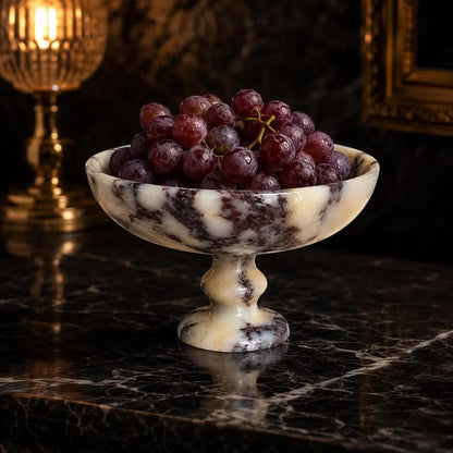 A marble pedestal bowl filled with dark red grapes, set on a polished black marble surface with a softly blurred warm-toned background.