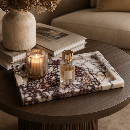 A rectangular marble-style tray with a white base and dark purple marbling sits on a round wooden table in a styled interior. The tray holds a lit candle in a ribbed glass holder, a small clear perfume bottle with gold detailing, and a gold chain.