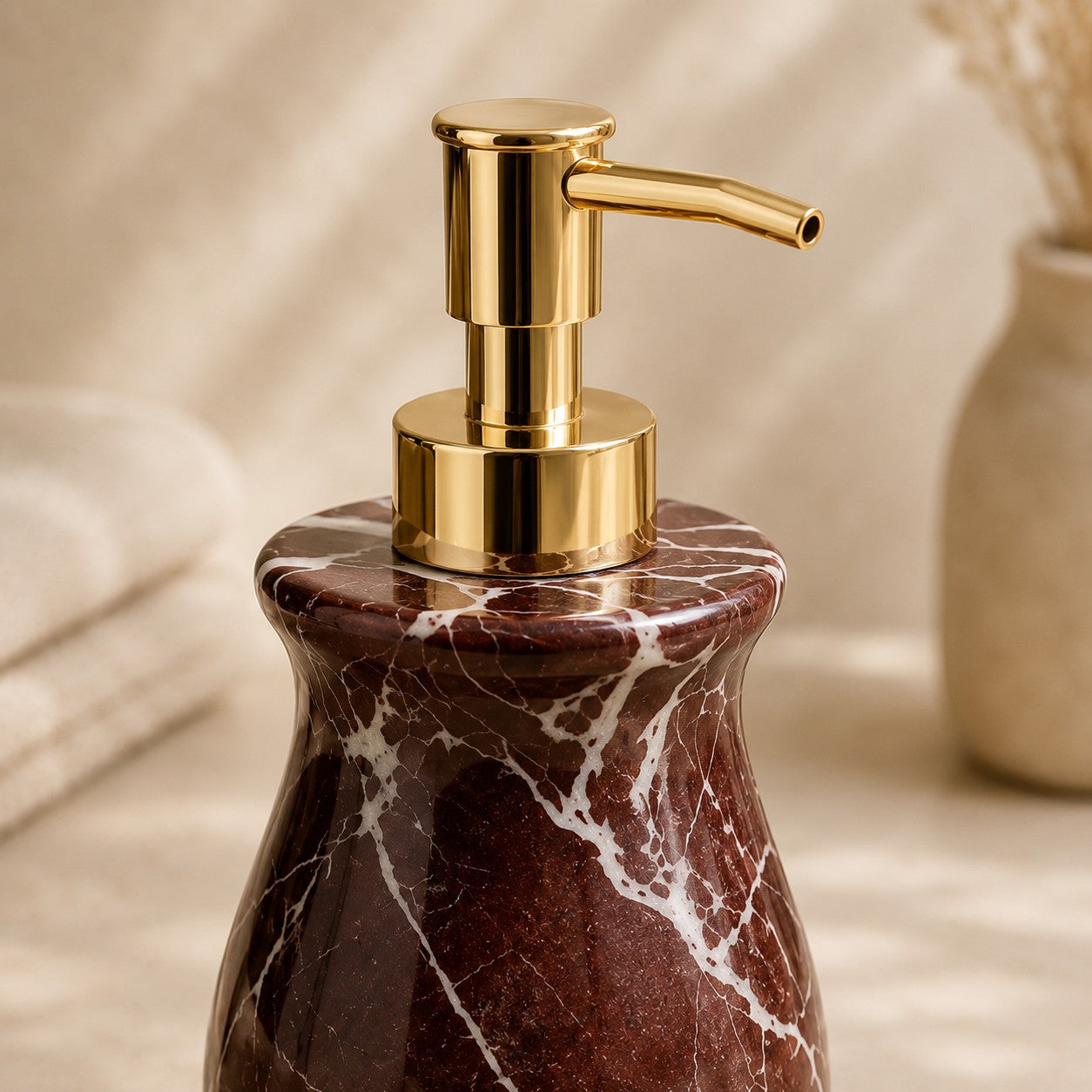 A close-up of a bathroom or vanity pump bottle with a polished gold-tone dispenser and a dark reddish-brown marble base featuring white veining, set against a softly blurred beige background with a folded towel and vase.