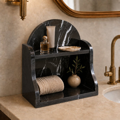 A black marble bathroom vanity organizer with two shelves, styled with a rolled beige towel, a small vase with greenery, a perfume bottle, and a soap dish, set on a beige marble countertop next to a brass faucet.