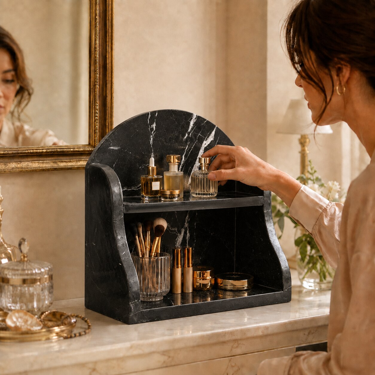 A woman in a beige robe organizes cosmetics on a black marble double-tier vanity organizer on a cream marble counter in a warm, elegant bathroom setting.