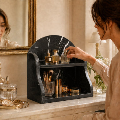 A woman in a beige robe organizes cosmetics on a black marble double-tier vanity organizer on a cream marble counter in a warm, elegant bathroom setting.