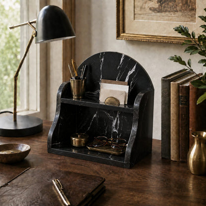 A black marble-look desk organizer with a curved back and two shelves, styled on a wooden desk with a brass lamp, books, glasses, and stationery in a warm indoor setting.