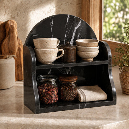 A black marble-look two-tier shelf organizer styled on a kitchen counter, holding stacked ceramic mugs, a small lidded jar, and glass jars with dried herbs and beans.