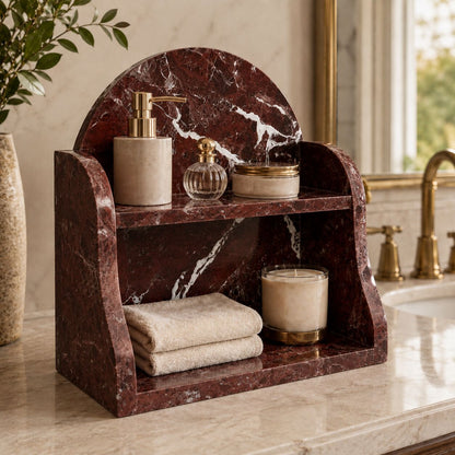 A two-tier bathroom vanity organizer made of dark reddish-brown marble, styled with beige soap dispensers, a small perfume bottle, a jar, and folded towels on a marble countertop.