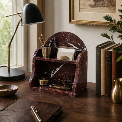 A dark red and white marble desk organizer with a curved back, styled on a wooden desk with a black desk lamp, books, and stationery.