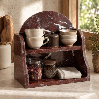 A two-tier marble kitchen organizer with a curved backrest, shown on a countertop with ceramic mugs, bowls, jars, and a folded cloth. The organizer has a dark reddish-brown marble finish with white veining.